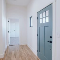A bright, minimalist hallway featuring light wood flooring, white walls, and a soft blue front door with glass panes, leading to an open interior space.